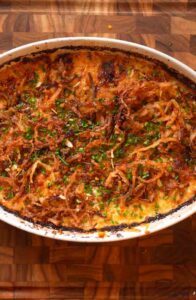 Golden brown scalloped potatoes baked in a white oval dish, topped with crispy fried onions, fresh herbs, and melted cheese on a wooden board.